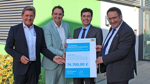 Vier Männer vor einem Gebäude mit einem symbolischen Geldscheck
