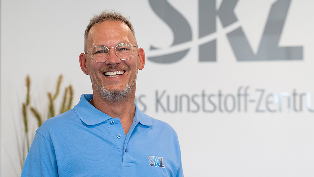 Ein Mann mit blauem Poloshirt lacht in die Kamera, im Hintergrund ist das Logo des SKZ